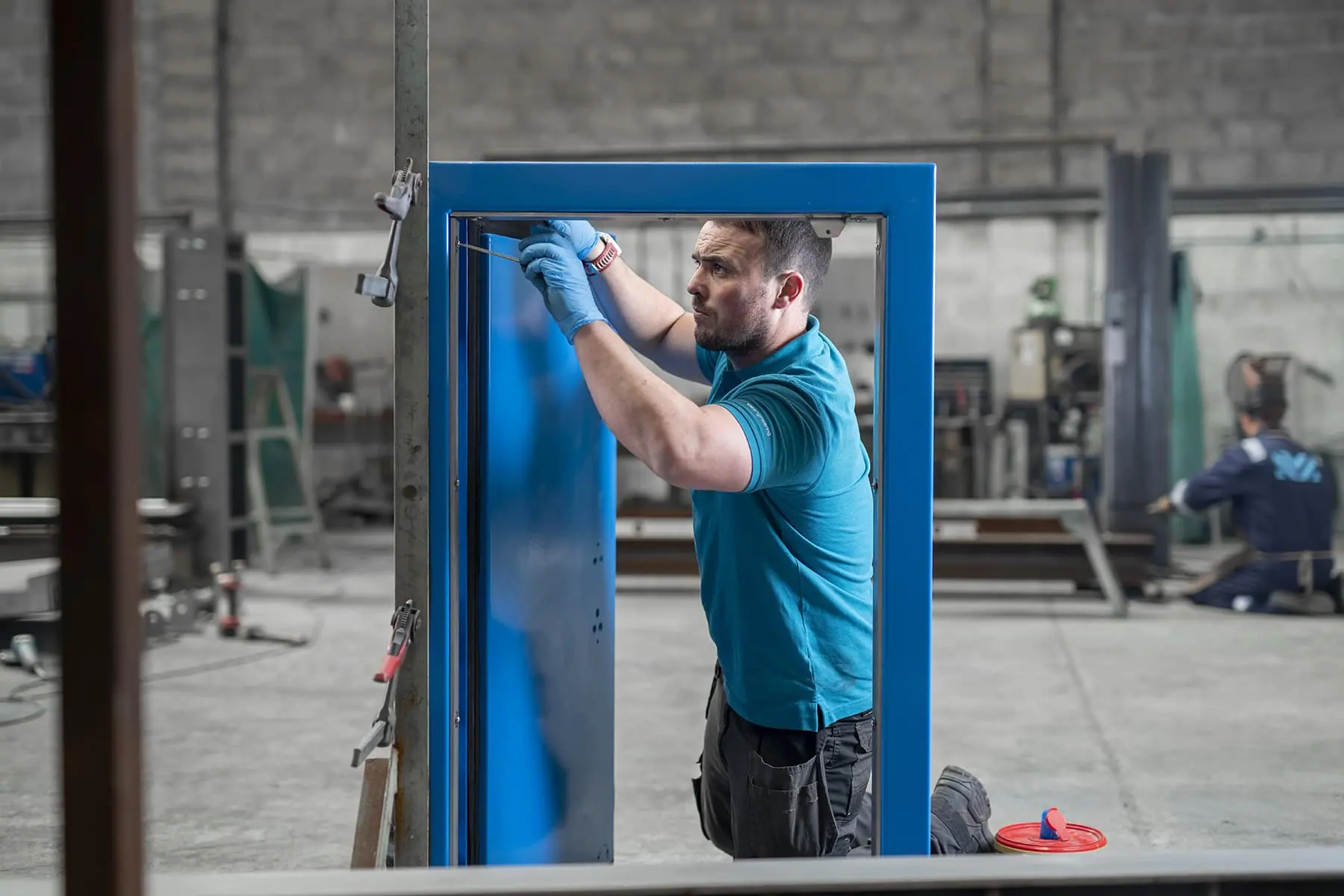 Fabricator in a teal polo shirt fitting a blue steel flood door within its frame in the workshop