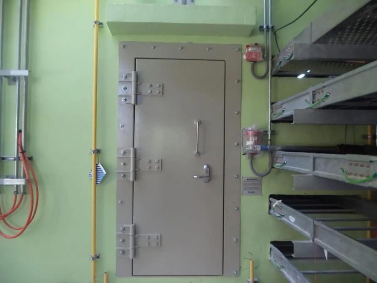 Single leaf beige blast door with bolted hinges on a green wall beside cable trays