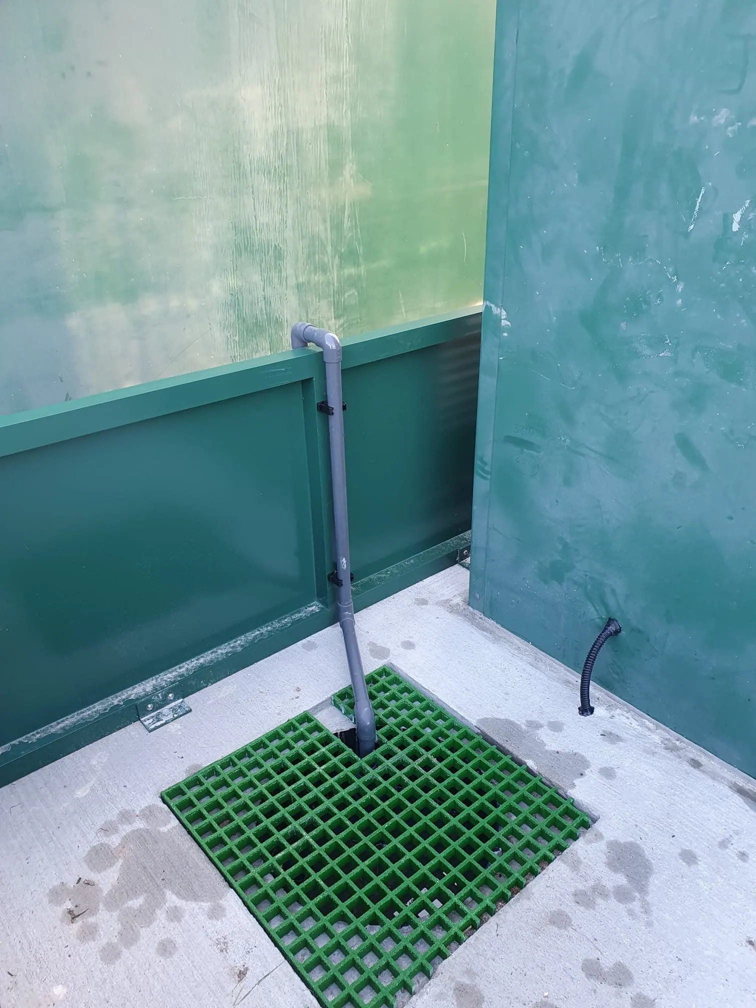 Inside a green flood barrier enclosure, a grey drain pipe running into a green grating