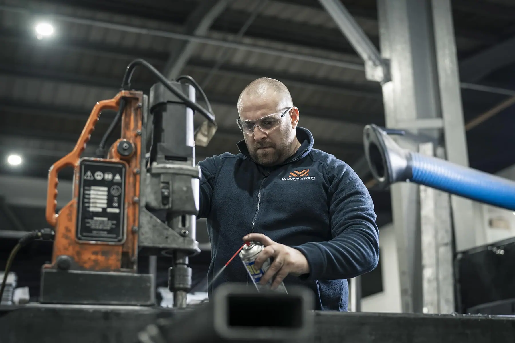 Engineer in safety glasses lubricating steel section beside a magnetic drill in the workshop