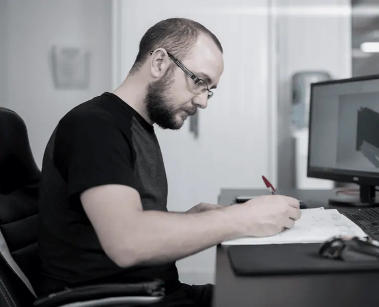 Design engineer sketching on paper at a desk beside a computer showing a 3D model