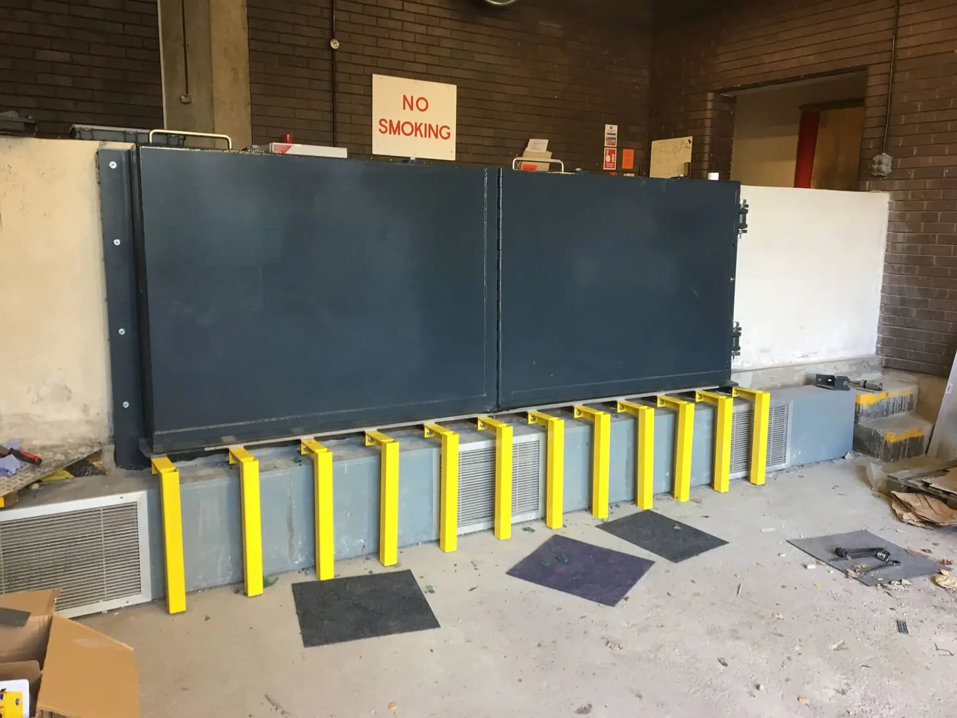 Hinged grey flood gate above bright yellow steel support brackets along a plinth