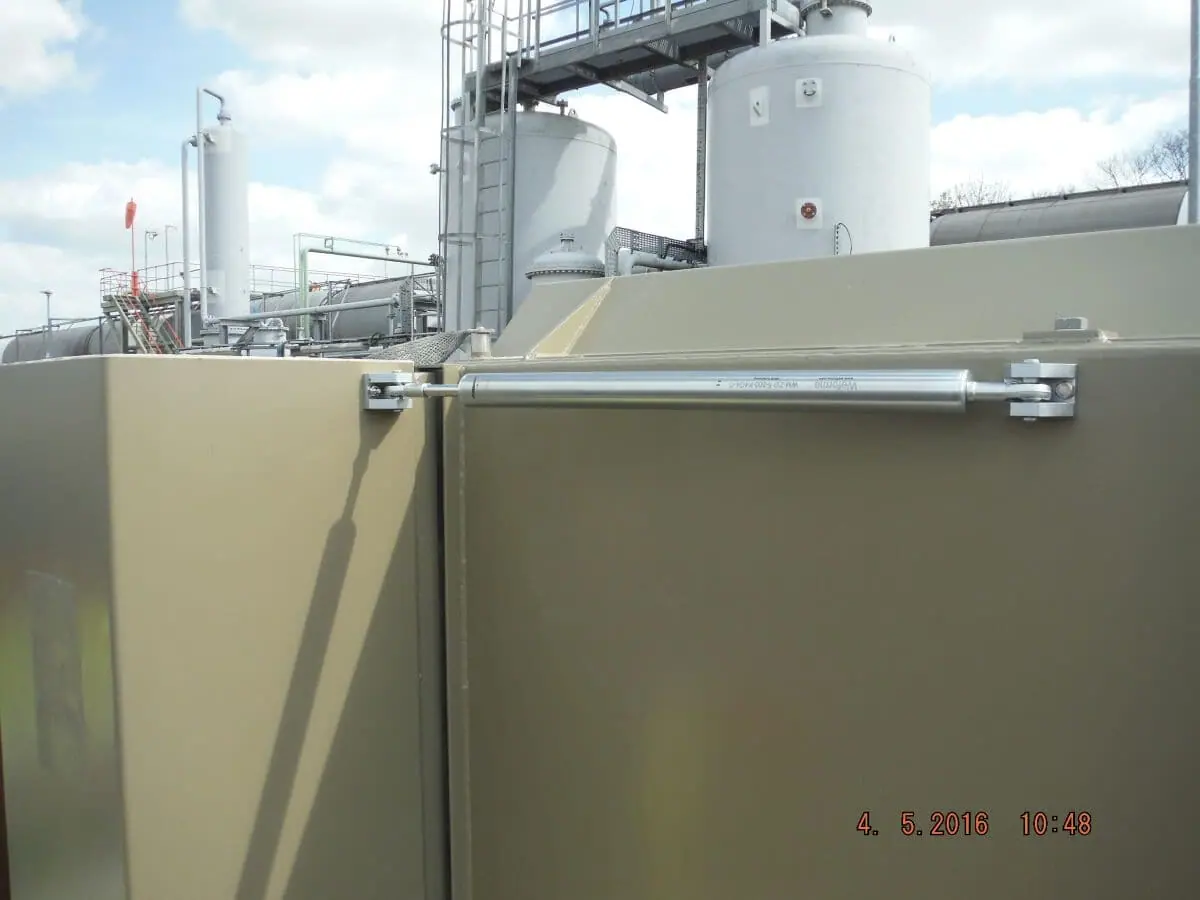 Stainless deceleration cylinder fitted to the dry side of a khaki flood gate beside industrial tanks