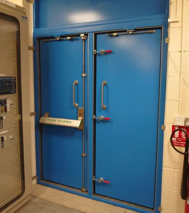 Blue double security flood door with push bar and red toggle clamps in a switch room