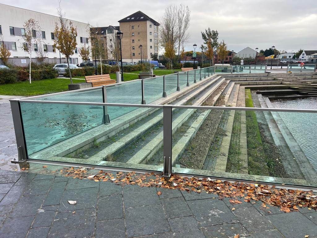 Glass flood wall running along stepped dock edge beside lawn and benches