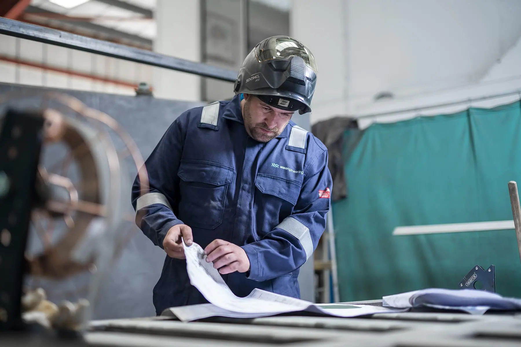 Engineer in MM Engineering overalls and welding helmet checking technical drawings in the workshop
