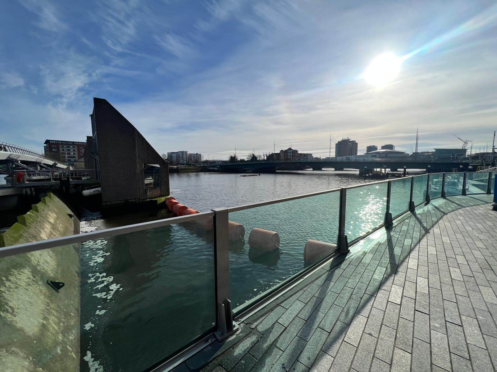 Glass flood wall with steel posts overlooking orange buoys, river and bridge in low sun