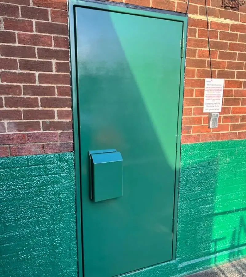 Green security flood door with letterbox set in a red brick wall with keypad