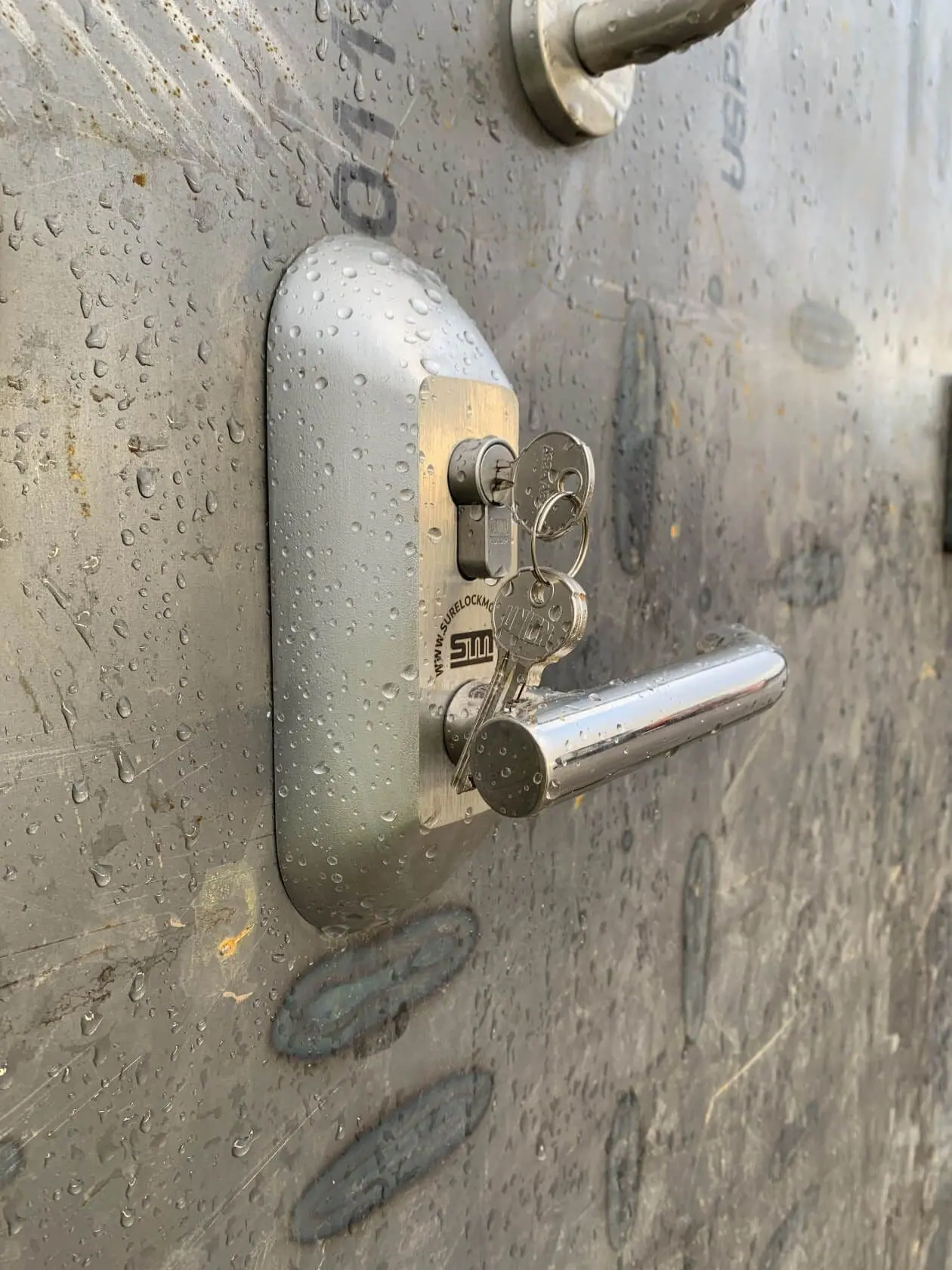 Stainless steel lock and lever handle with keys on a rain-covered blast test door
