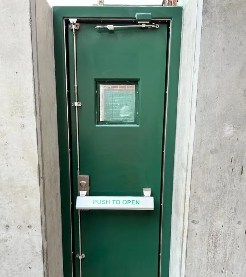 Green single flood door with vision panel and push bar between concrete walls