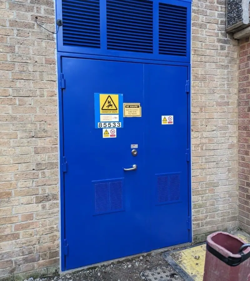 Blue security double door with louvred panels and warning signs in a brick substation wall
