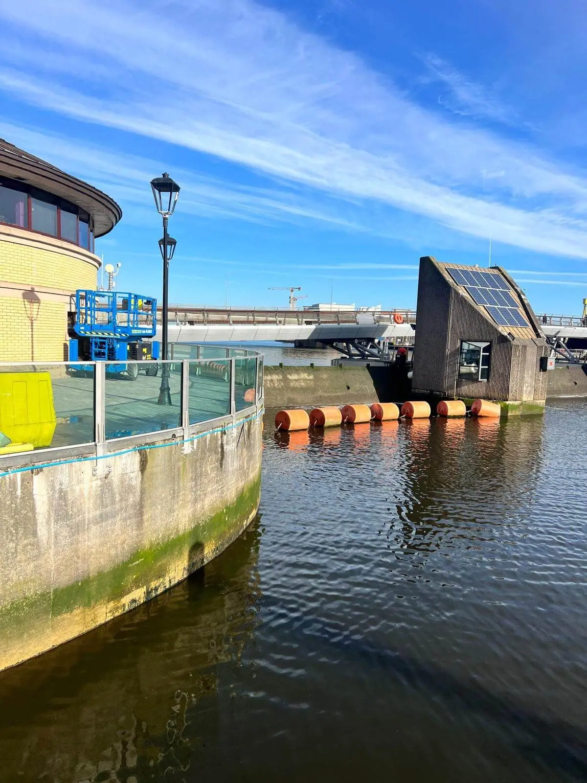 Curved glass flood wall on a quayside with orange buoys and a solar-panelled building