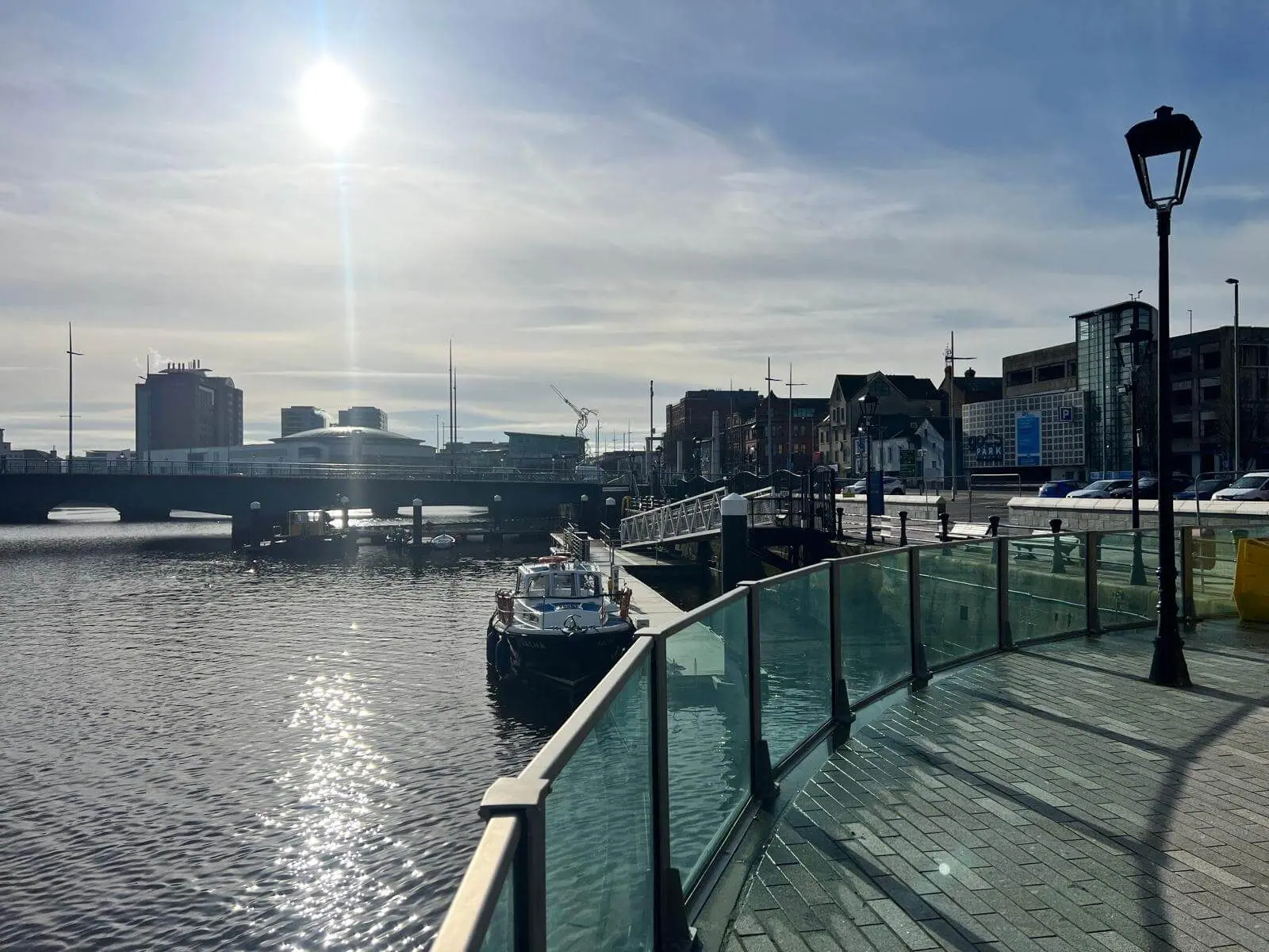 Curving glass flood wall along a paved waterfront with a moored boat and bridge