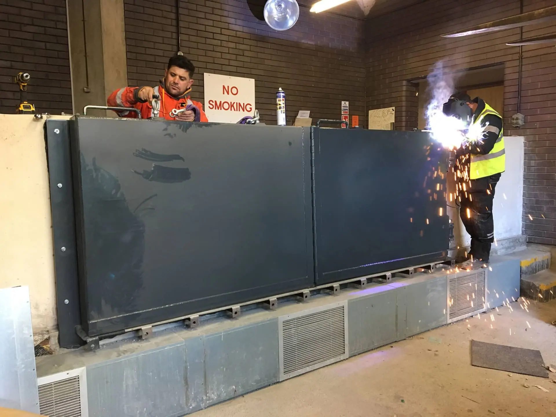Engineers welding and rigging a dark grey flood barrier installed inside a brick building