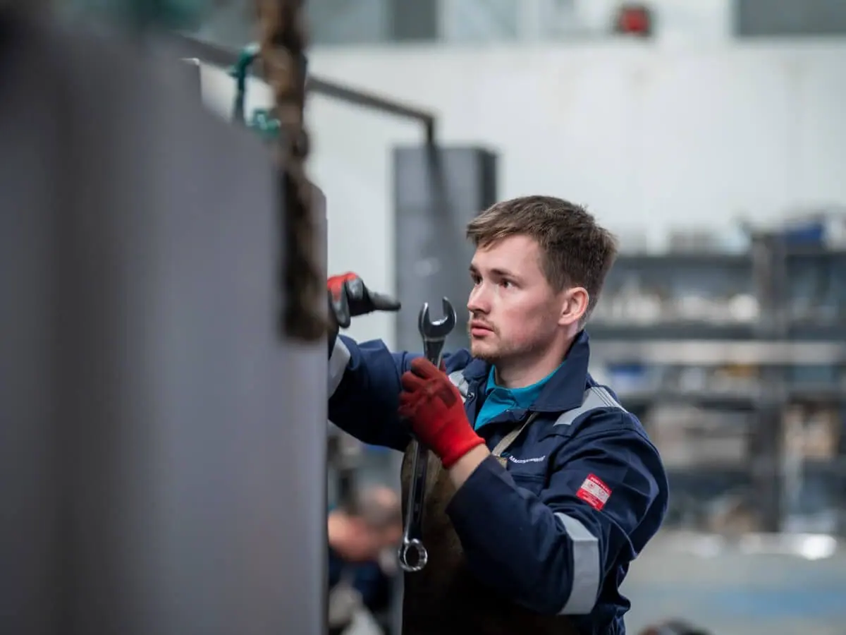 Fabricator in navy overalls and red gloves holding a spanner beside a steel door in the workshop