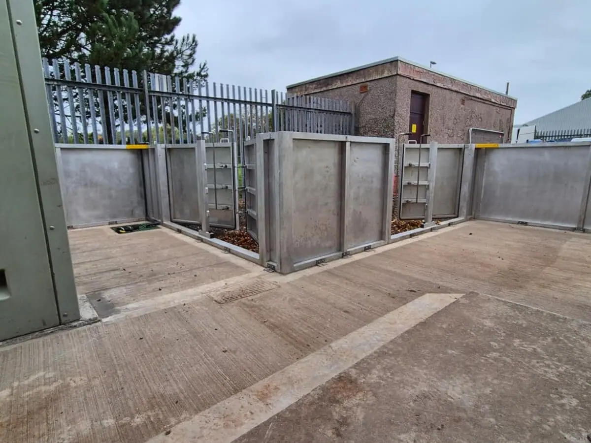 Grey metal flood barrier panels with access gates surrounding electrical equipment on a concrete yard