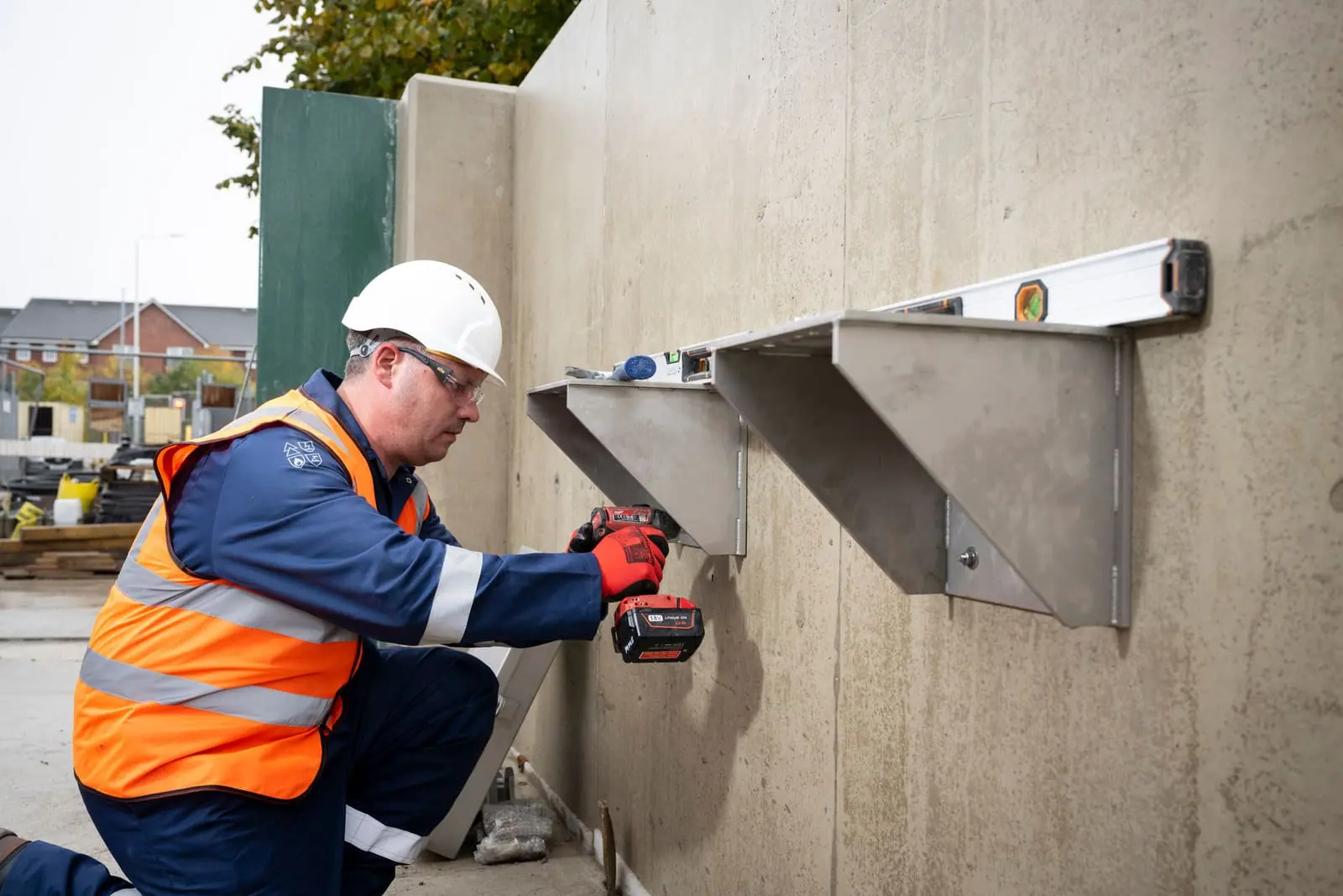 Installer kneeling to drill stainless steel mounting brackets into a concrete wall, using a spirit level