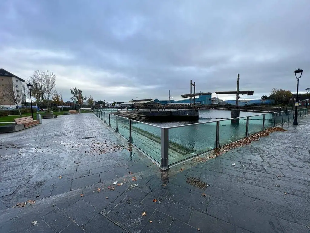 Glass flood wall enclosing a paved dockside beside a harbour basin and lifting bridge