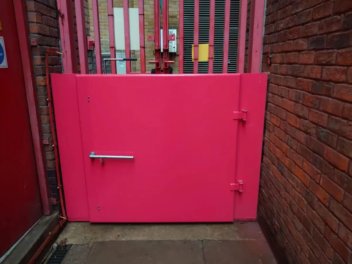 Pink steel flood door with handle and hinges fitted in a narrow brick alcove