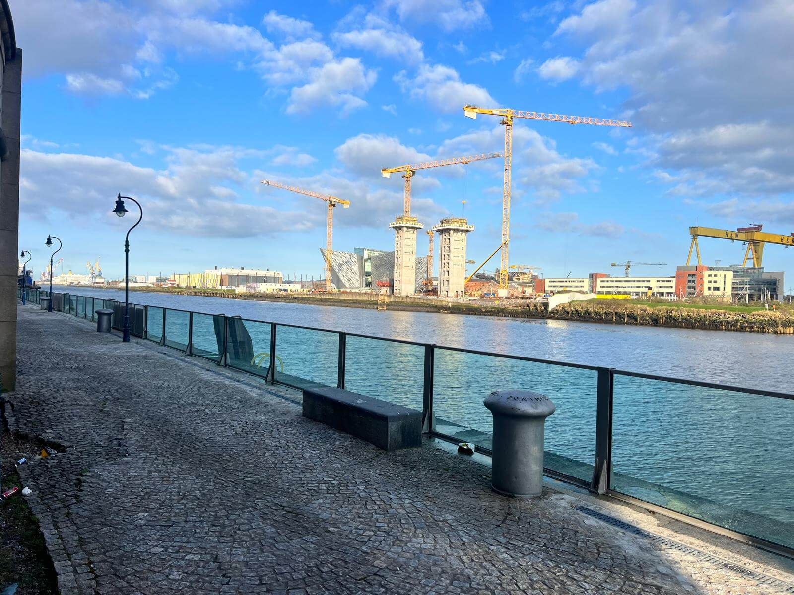 Glass flood wall along a cobbled riverside walkway with cranes across the water