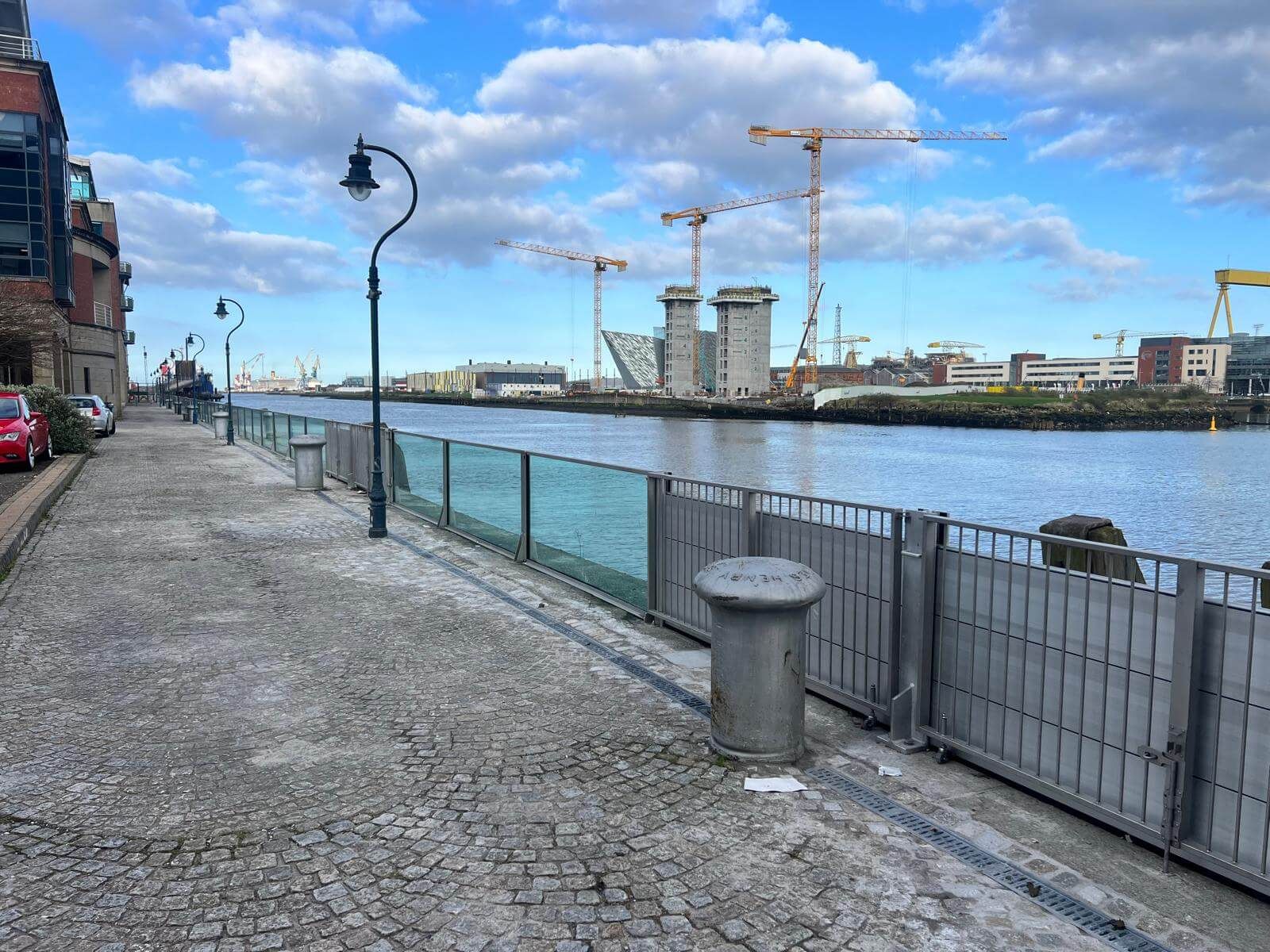 Flood barrier with glass panels and sliding steel gate along a riverside walkway with cranes beyond