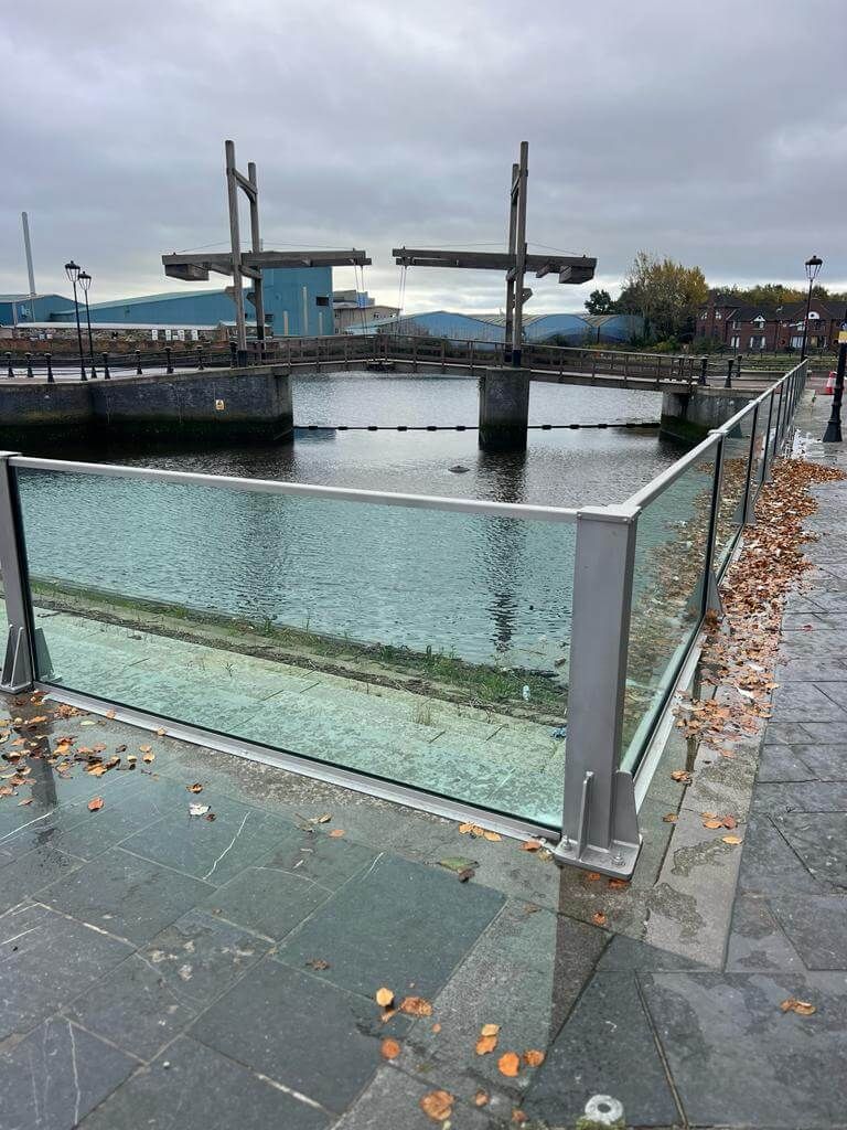 Close view of glass flood wall panels with steel posts above a dock, autumn leaves on paving