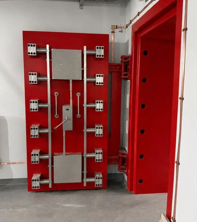 Red steel blast door with multiple locking bolts and central handle fitted in a concrete frame
